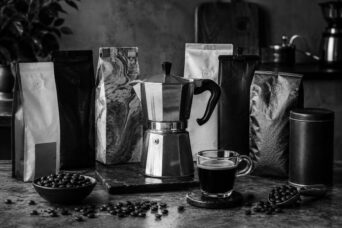 Moka pot and glass espresso cup surrounded by coffee bags in various textures, loose beans, a ceramic bowl, and a metal tin on a dark stone surface, shot in moody black and white.