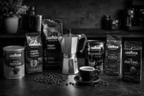 Several Lavazza coffee bags and a tin arranged around a stovetop moka pot and espresso cup on a dark stone surface, with scattered coffee beans and a blurred kitchen background, shot in black and white.