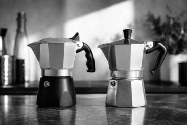 Two moka pots on a countertop: a black Bialetti Brikka on the left and a silver Moka Express on the right, bathed in soft natural light.