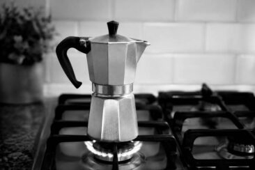 A moka pot sits on a gas stovetop, with a blurred background of light-colored tiles and a hint of foliage in a container.