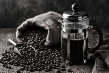 A French press filled with dark coffee sits next to a burlap sack spilling dark roast coffee beans, with a metal scoop partially filled with beans on a wooden surface.