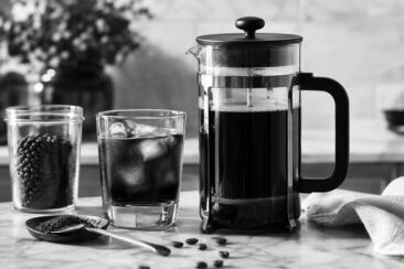 A French press with brewed coffee, a glass of iced coffee with ice cubes, a jar of coffee beans, and a spoon of ground coffee on a marble countertop.