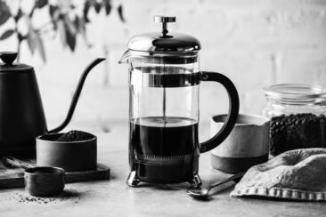 A black and white image of a coffee setup featuring a French press, a spouted kettle, a container of ground coffee, and a jar of coffee beans on a wooden surface.