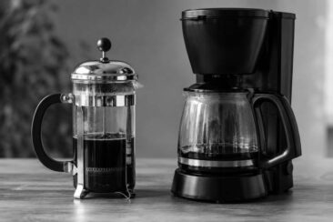 A French press and a drip coffee maker are placed side by side on a wooden surface, both containing dark coffee and highlighting various shapes and textures.