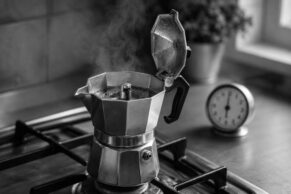 Stovetop moka pot brewing on a gas stove with steam rising from the open lid, a round clock and potted plant softly blurred in the background, captured in moody black and white.
