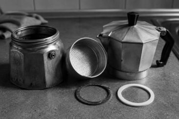 Black and white photo of a Moka pot and a rubber gasket ring placed beside it on a textured surface.