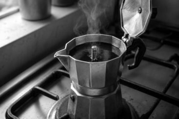 Aluminum moka pot brewing on a gas stove with steam rising from the open lid, captured in black and white with a moody, atmospheric feel.