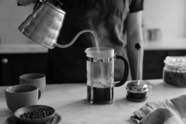 A person pours hot water from a metallic kettle into a French press filled with coffee grounds, surrounded by coffee cups and beans on a kitchen countertop.