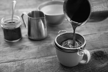 A metallic jug pours dark coffee into a strainer above a white cup, with a glass jar of coffee grounds and a metal creamer in the scene.