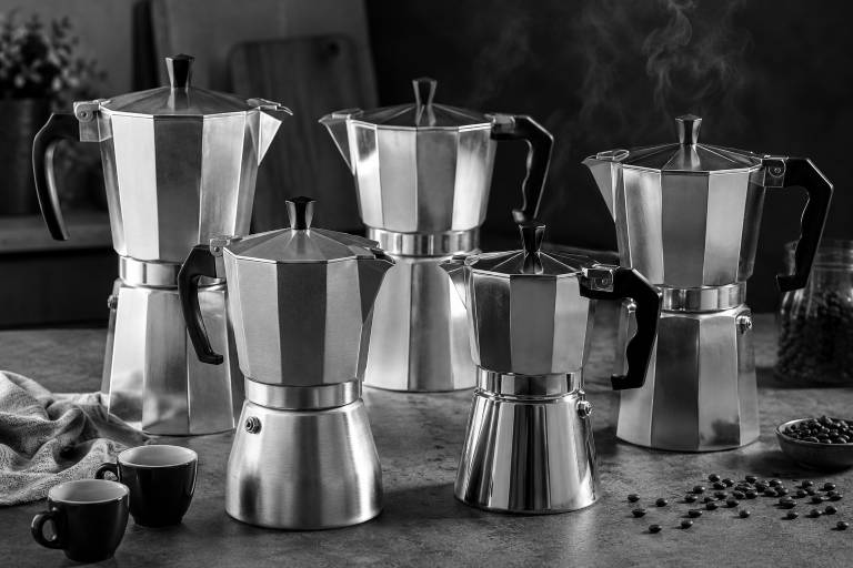 A collection of five moka pots in metal, with steam rising from one, two black coffee cups, and scattered coffee beans on a textured surface.