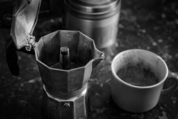 A moka pot with an open lid beside a stained white coffee cup on a dark surface.