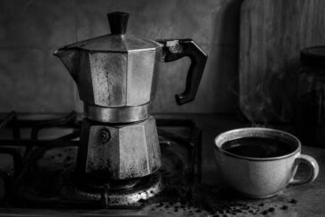 A worn stovetop espresso maker sits on a gas stove next to a steaming cup of dark coffee, with coffee grounds scattered nearby.