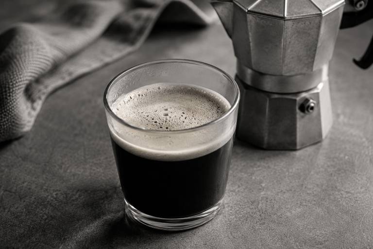 A clear glass of dark coffee with crema next to a metallic moka pot on a gray surface.