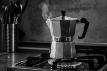 Polished aluminum moka pot steaming on a lit gas burner, with kitchen utensils blurred in the background and a moody black-and-white tone.