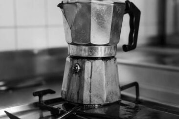 Well-worn aluminum moka pot sitting on a gas stovetop burner, with coffee stains and a matte patina, captured in black and white with soft background blur.