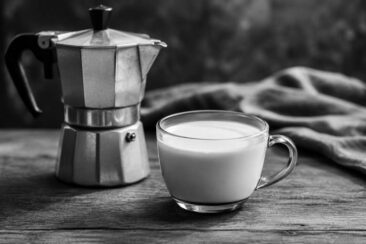 A vintage moka pot beside a glass cup filled with milk, set on a wooden tabletop, with a soft gray cloth in the background.