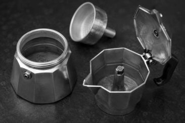 Disassembled aluminum moka pot with lid open, filter basket, and bottom chamber separated on a dark slate surface, shot in black and white.