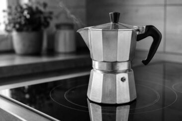 Polished aluminum moka pot with a black handle releasing a wisp of steam on a black glass ceramic stovetop, with a blurred potted plant and canister in the background.