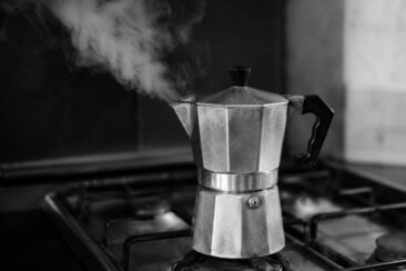 Polished aluminum moka pot on a gas stovetop releasing a wisp of steam, captured in black and white with a softly blurred dark kitchen background.