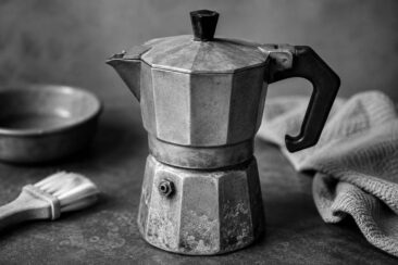 Worn aluminum moka pot on a dark stone surface with a small brush, ceramic bowl, and folded linen cloth in a moody black-and-white close-up.