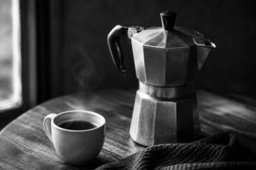 A black and white image of a stovetop espresso maker beside a steaming cup of coffee on a wooden table, with a folded dark cloth nearby.
