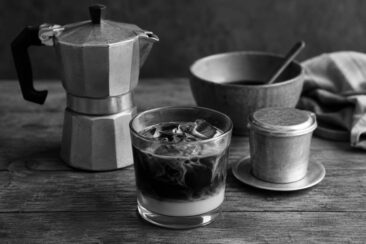 Black and white vignette of an iced coffee in a rocks glass with swirling cream, a moka pot, a Vietnamese phin filter, a ceramic bowl, and a linen cloth on a rustic wooden surface.