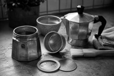 Disassembled moka pot with filter basket, gasket, and perforated plate laid out on a wet stone surface beside a pastry brush, linen cloth, and ceramic bowl, shot in black and white.