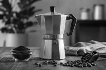 Aluminum moka pot on a dark wooden surface with a glass bowl of ground coffee, scattered coffee beans, a metal scoop, and a linen cloth, photographed in black and white with soft bokeh background.