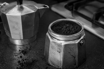 A stainless steel moka pot next to a container filled with ground coffee. The scene is in black and white, with coffee grounds spilled around the container, showcasing a kitchen setting with a gas stove in the background.