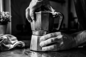 Two hands assembling a well-worn moka pot on a dark countertop, with scattered coffee grounds, a folded linen cloth, and soft background blur in moody black and white.