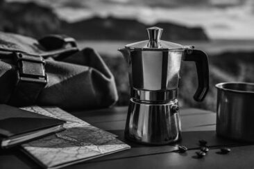 A silver moka pot beside a metallic cup and scattered coffee beans on a wooden table, with a dark backpack and a black notebook on a map. Blurred hills and water are in the background.