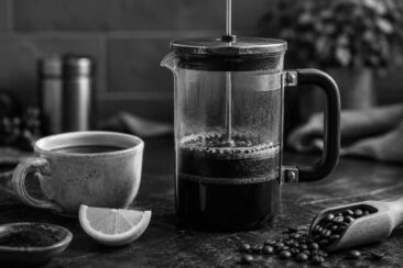 A French press with coffee, a speckled ceramic cup of coffee, and a wedge of lemon on a dark tabletop, accompanied by scattered coffee beans.